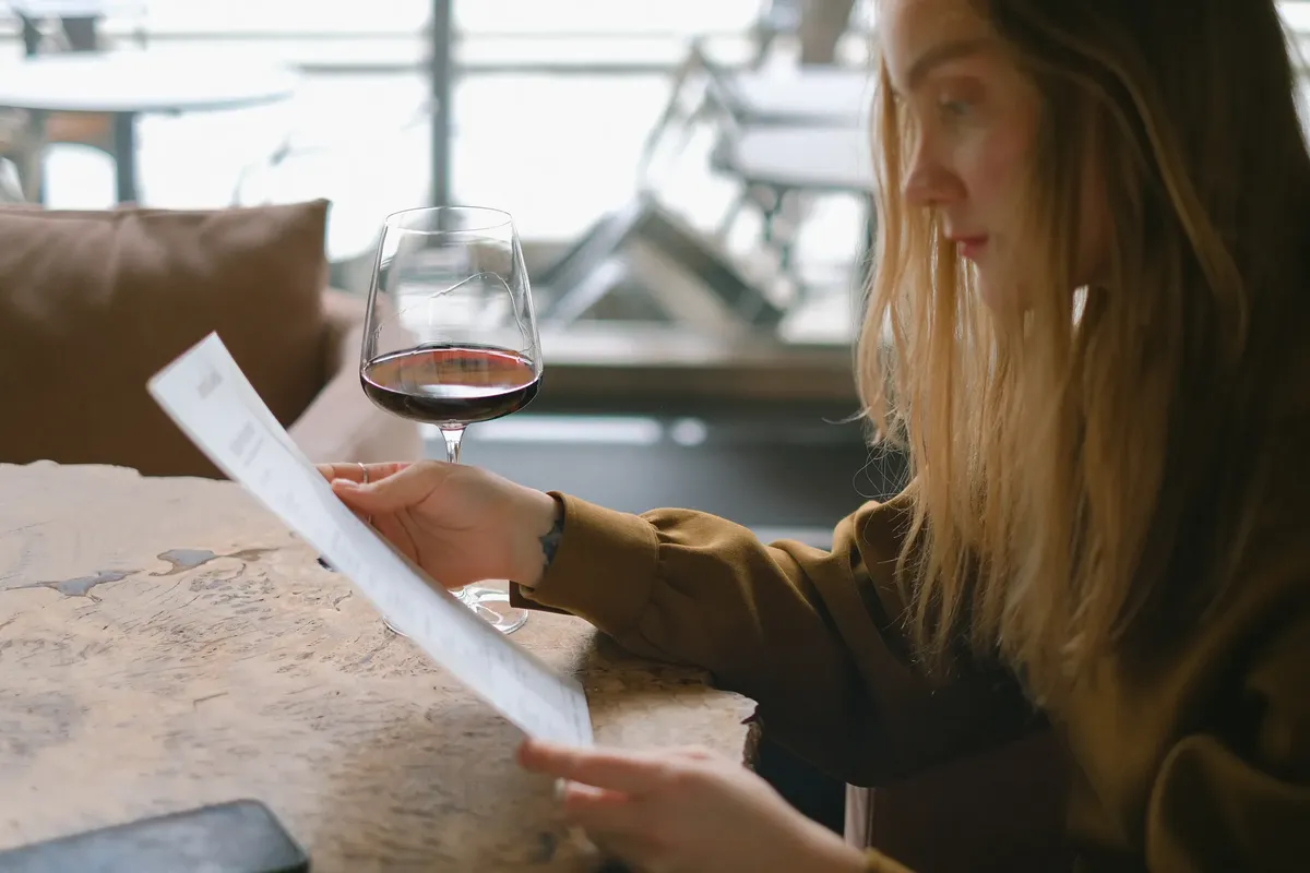 A woman reads a menu while holding a glass of red wine at a restaurant.