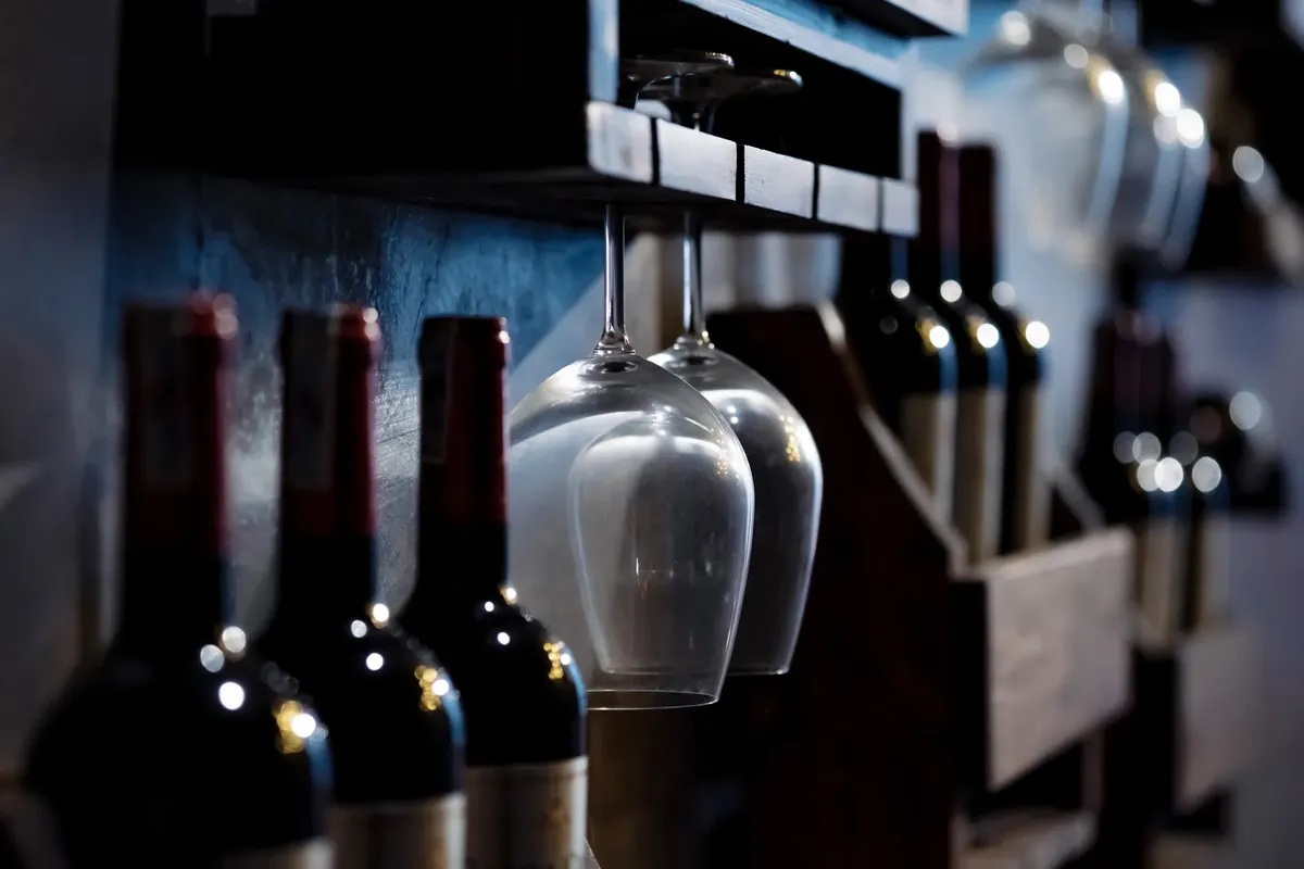 Close-up of wine bottles and glasses on wooden shelves in a dimly lit cellar, creating a cozy ambiance.